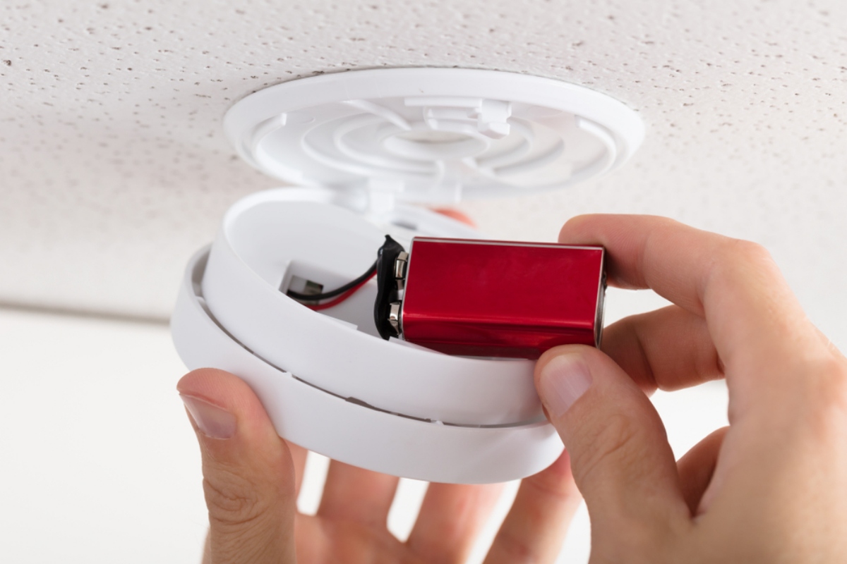 Person inserting a red battery into a white smoke detector housing on the ceiling. When finished, the cover closes back into place.
