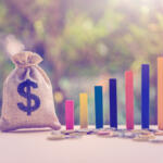 Money bag with a dollar sign beside a rising multicolored bar chart and coins, symbolizing financial growth and savings growth.