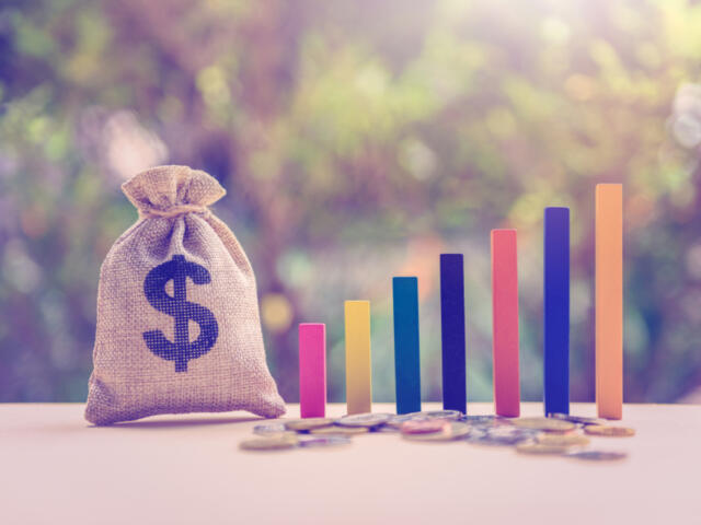 Money bag with a dollar sign beside a rising multicolored bar chart and coins, symbolizing financial growth and savings growth.