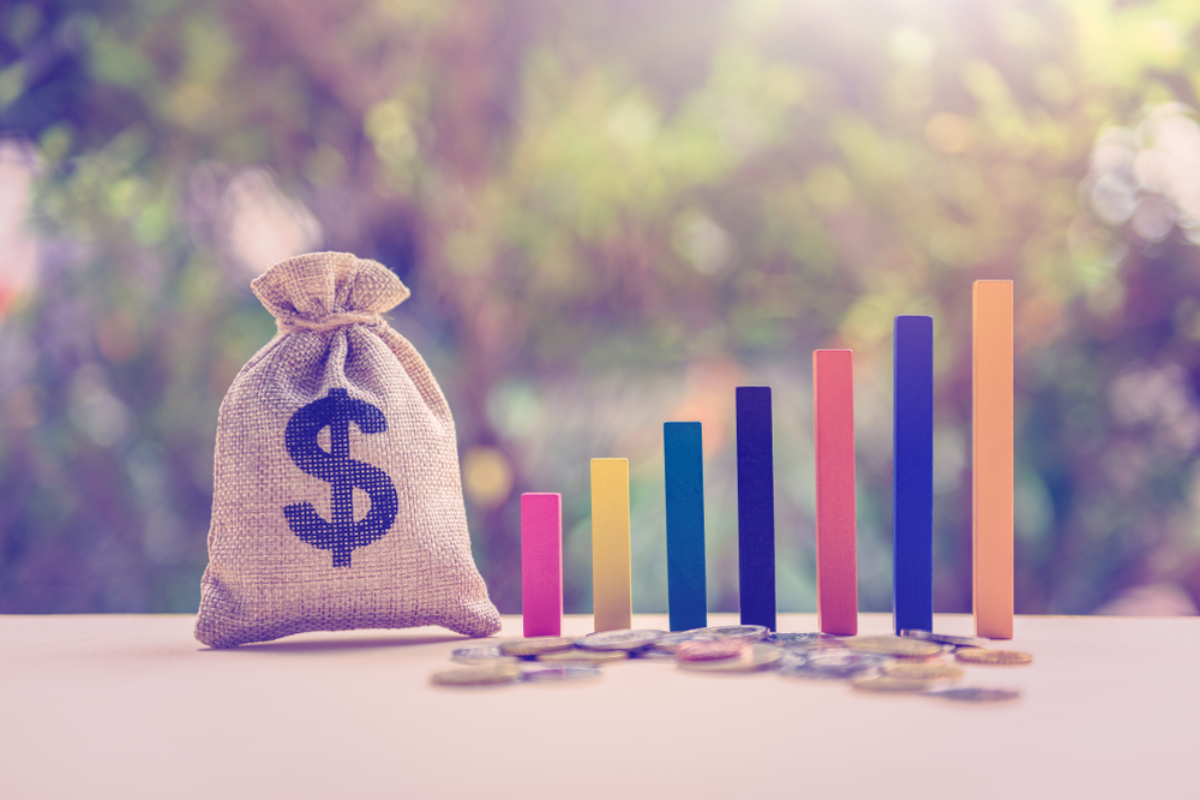 Money bag with a dollar sign beside a rising multicolored bar chart and coins, symbolizing financial growth and savings growth.