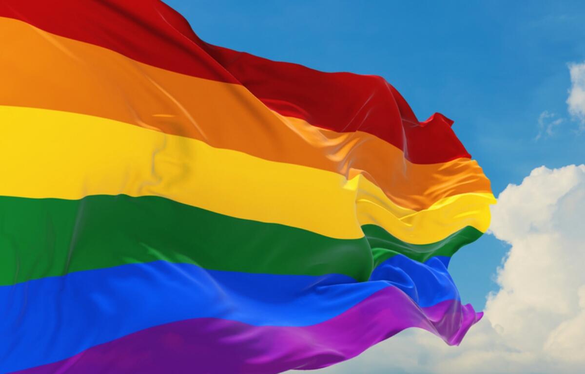 Rainbow Pride flag waving in the wind against a blue sky with white clouds behind it.