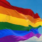 Rainbow Pride flag waving in the wind against a blue sky with white clouds behind it.