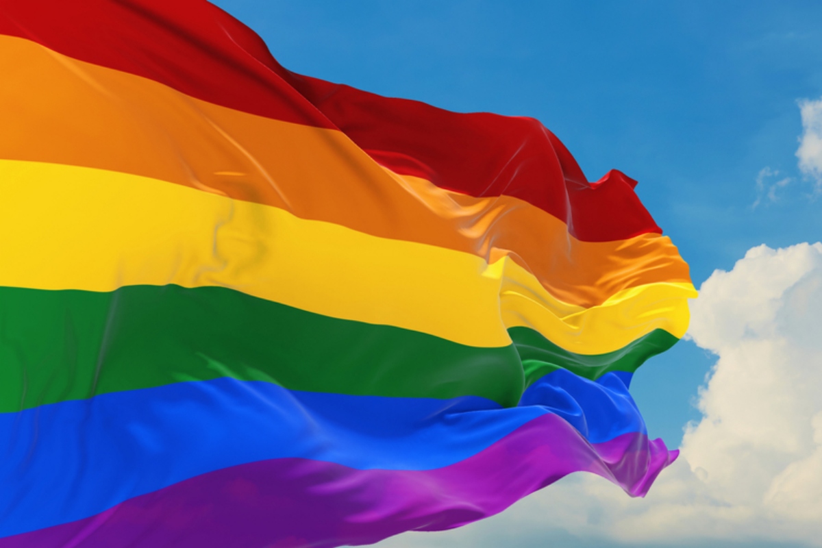 Rainbow Pride flag waving in the wind against a blue sky with white clouds behind it.