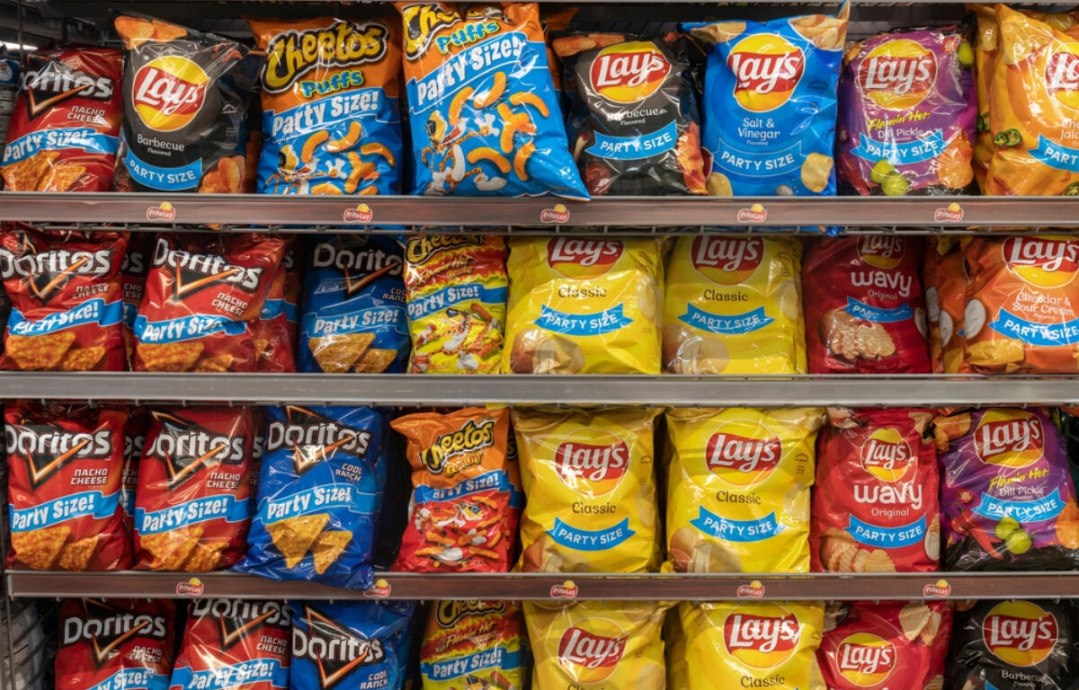 Shelf of assorted party-size snack bags on supermarket shelves, including Lay's, Doritos, and Cheetos-style brands.