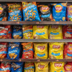 Shelf of assorted party-size snack bags on supermarket shelves, including Lay's, Doritos, and Cheetos-style brands.