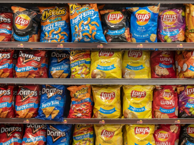 Shelf of assorted party-size snack bags on supermarket shelves, including Lay's, Doritos, and Cheetos-style brands.