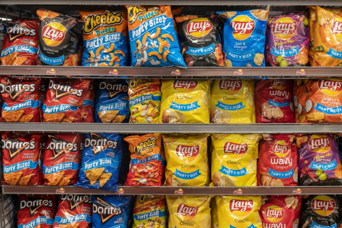 Shelf of assorted party-size snack bags on supermarket shelves, including Lay's, Doritos, and Cheetos-style brands.