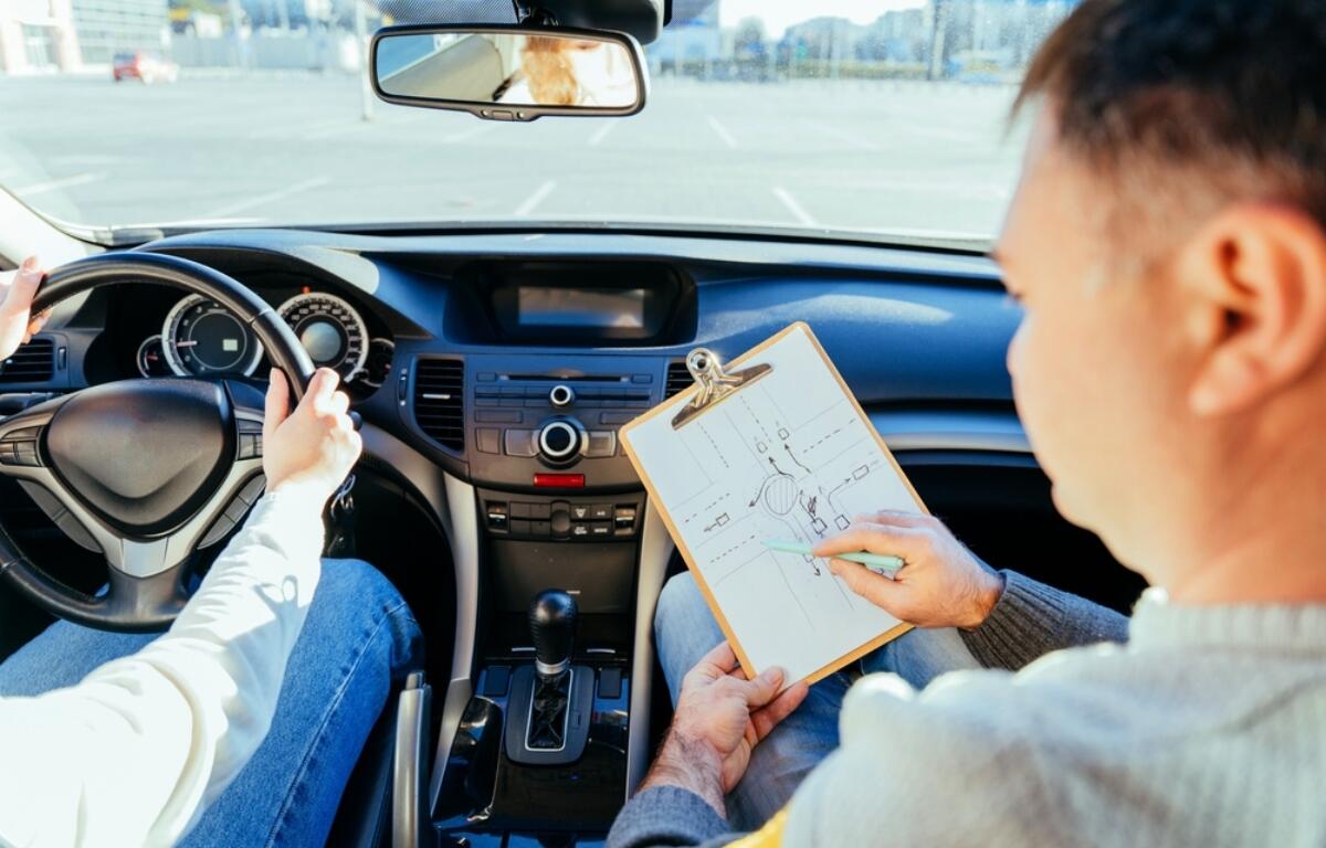 Driver grips the steering wheel while an instructor sits beside, showing a clipboard with a diagram inside the car