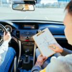 Driver grips the steering wheel while an instructor sits beside, showing a clipboard with a diagram inside the car