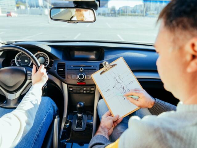 Driver grips the steering wheel while an instructor sits beside, showing a clipboard with a diagram inside the car