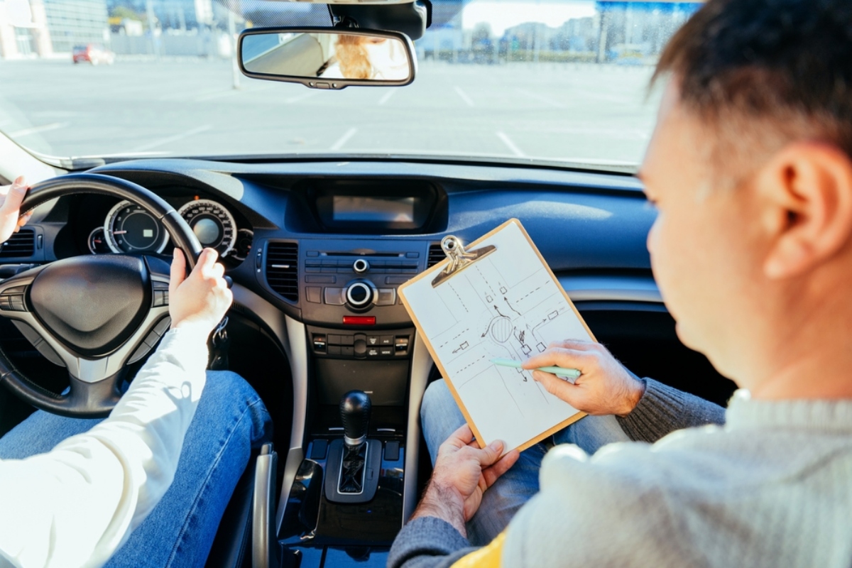 Driver grips the steering wheel while an instructor sits beside, showing a clipboard with a diagram inside the car