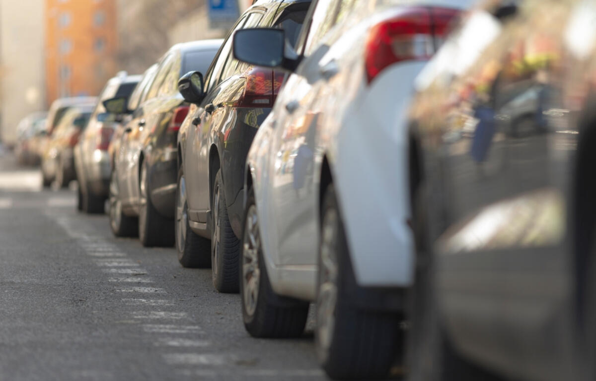 Row of cars parked along a city street, viewed from the curb with buildings in the background.