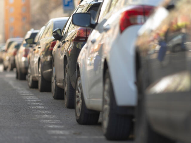 Row of cars parked along a city street, viewed from the curb with buildings in the background.