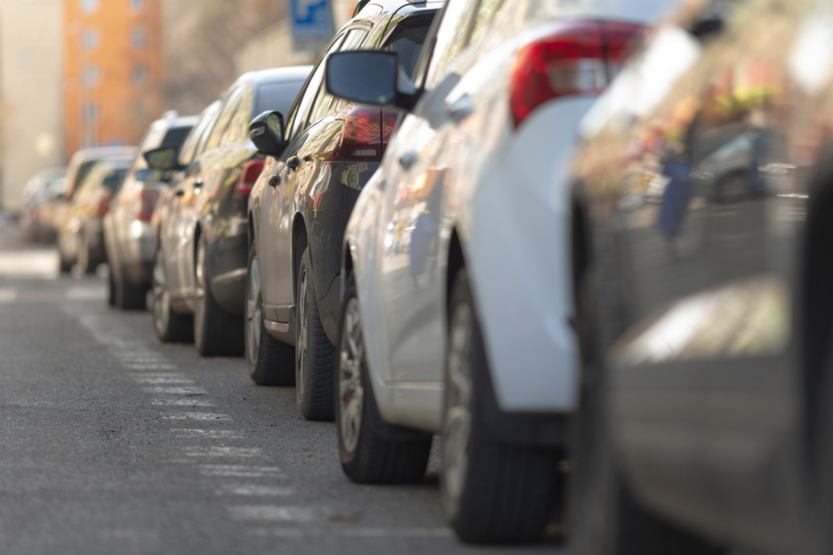 Row of cars parked along a city street, viewed from the curb with buildings in the background.