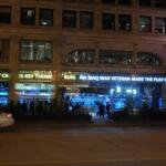 A small crowd gathers outside the ABC Channel 7 studio in downtown Chicago to watch a live broadcast of the 2008 presidential debates through the station’s street‑facing window. Pedestrians pause along State Street as the brightly lit newsroom displays the televised event, creating a moment of shared public viewing in the city’s nighttime streetscape. Chicago, IL, USA October 02, 2008