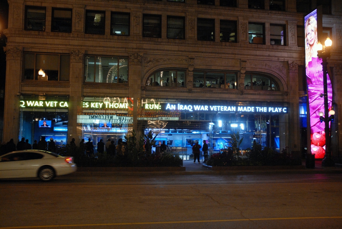 A small crowd gathers outside the ABC Channel 7 studio in downtown Chicago to watch a live broadcast of the 2008 presidential debates through the station’s street‑facing window. Pedestrians pause along State Street as the brightly lit newsroom displays the televised event, creating a moment of shared public viewing in the city’s nighttime streetscape. Chicago, IL, USA October 02, 2008