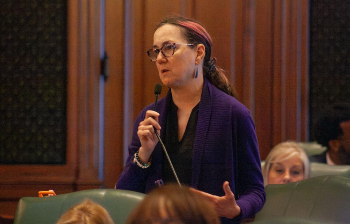 Woman with glasses and a purple cardigan speaking into a microphone in a formal meeting room, gesturing with her other hand.