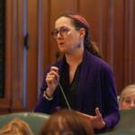 Woman with glasses and a purple cardigan speaking into a microphone in a formal meeting room, gesturing with her other hand.