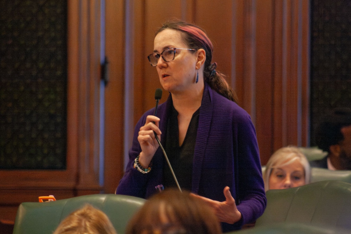 Woman with glasses and a purple cardigan speaking into a microphone in a formal meeting room, gesturing with her other hand.
