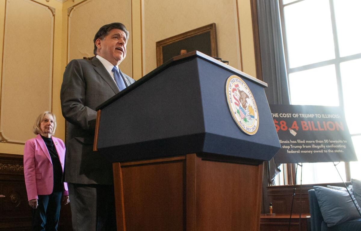 Man in a gray suit speaks from a blue podium with a government seal; a woman in a pink blazer stands behind him in a wood-paneled room.