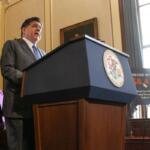 Man in a gray suit speaks from a blue podium with a government seal; a woman in a pink blazer stands behind him in a wood-paneled room.