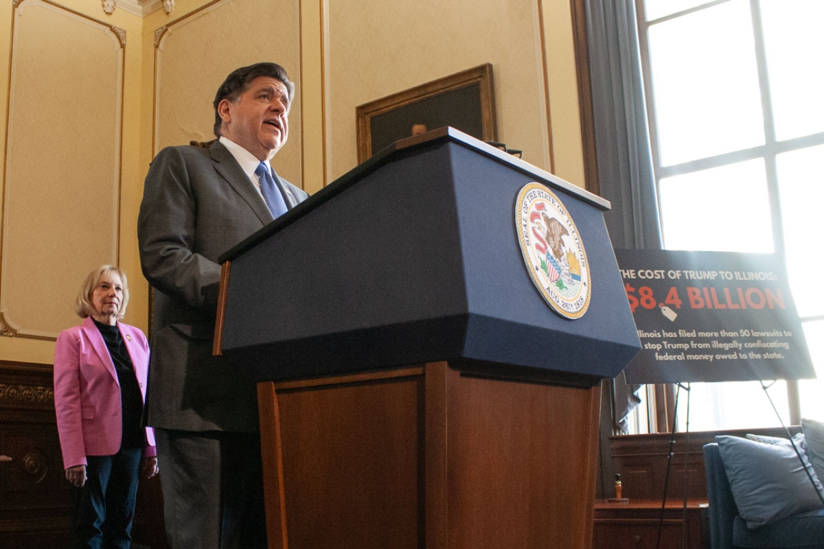 Man in a gray suit speaks from a blue podium with a government seal; a woman in a pink blazer stands behind him in a wood-paneled room.