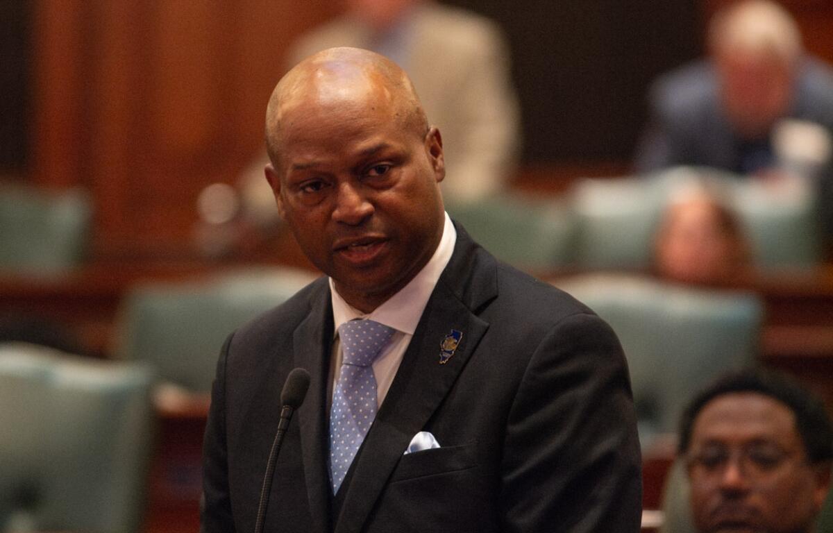 Man in a dark suit and light blue polka-dot tie speaks at a microphone in a formal chamber.