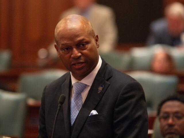 Man in a dark suit and light blue polka-dot tie speaks at a microphone in a formal chamber.