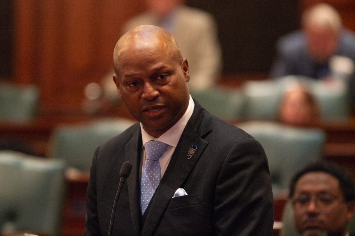 Man in a dark suit and light blue polka-dot tie speaks at a microphone in a formal chamber.