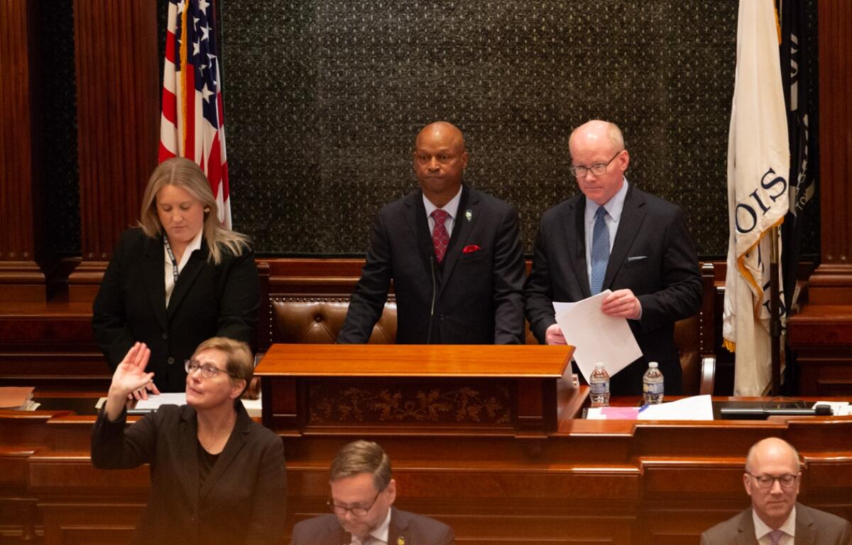 Man in a dark suit at a wooden podium addressing a formal session, flanked by two colleagues in a legislative chamber with flags behind them.