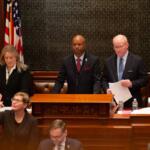 Man in a dark suit at a wooden podium addressing a formal session, flanked by two colleagues in a legislative chamber with flags behind them.
