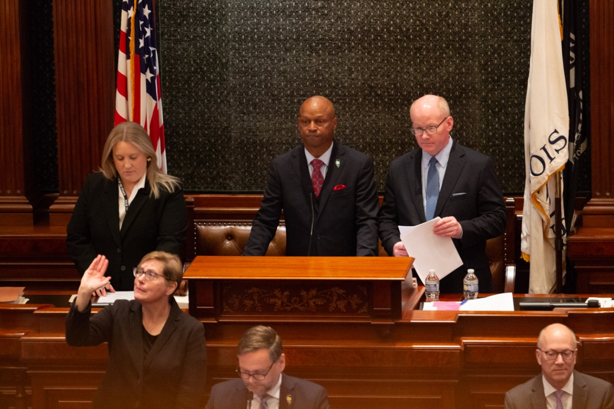 Man in a dark suit at a wooden podium addressing a formal session, flanked by two colleagues in a legislative chamber with flags behind them.