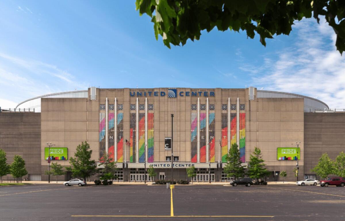 Front view of the United Center exterior with tall colorful banners along the facade, a spacious parking lot, and green trees against a blue sky.