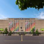 Front view of the United Center exterior with tall colorful banners along the facade, a spacious parking lot, and green trees against a blue sky.