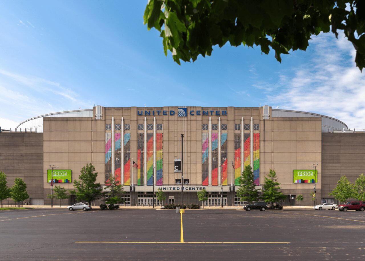 Front view of the United Center exterior with tall colorful banners along the facade, a spacious parking lot, and green trees against a blue sky.
