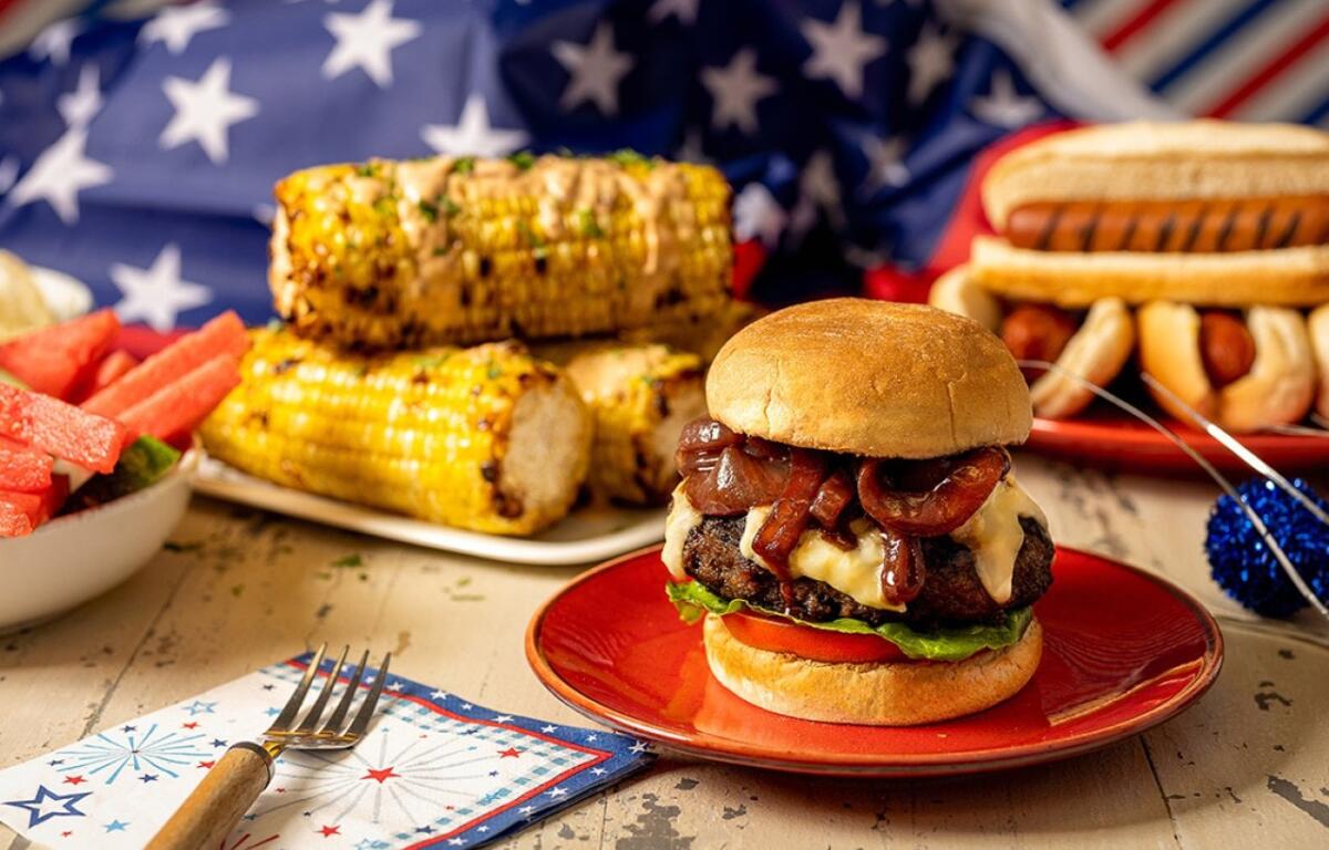 Cheeseburger with bacon and melted cheese on a bun, served on a red plate with corn on the cob and patriotic decorations in the background.