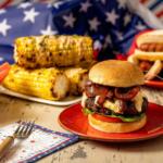 Cheeseburger with bacon and melted cheese on a bun, served on a red plate with corn on the cob and patriotic decorations in the background.
