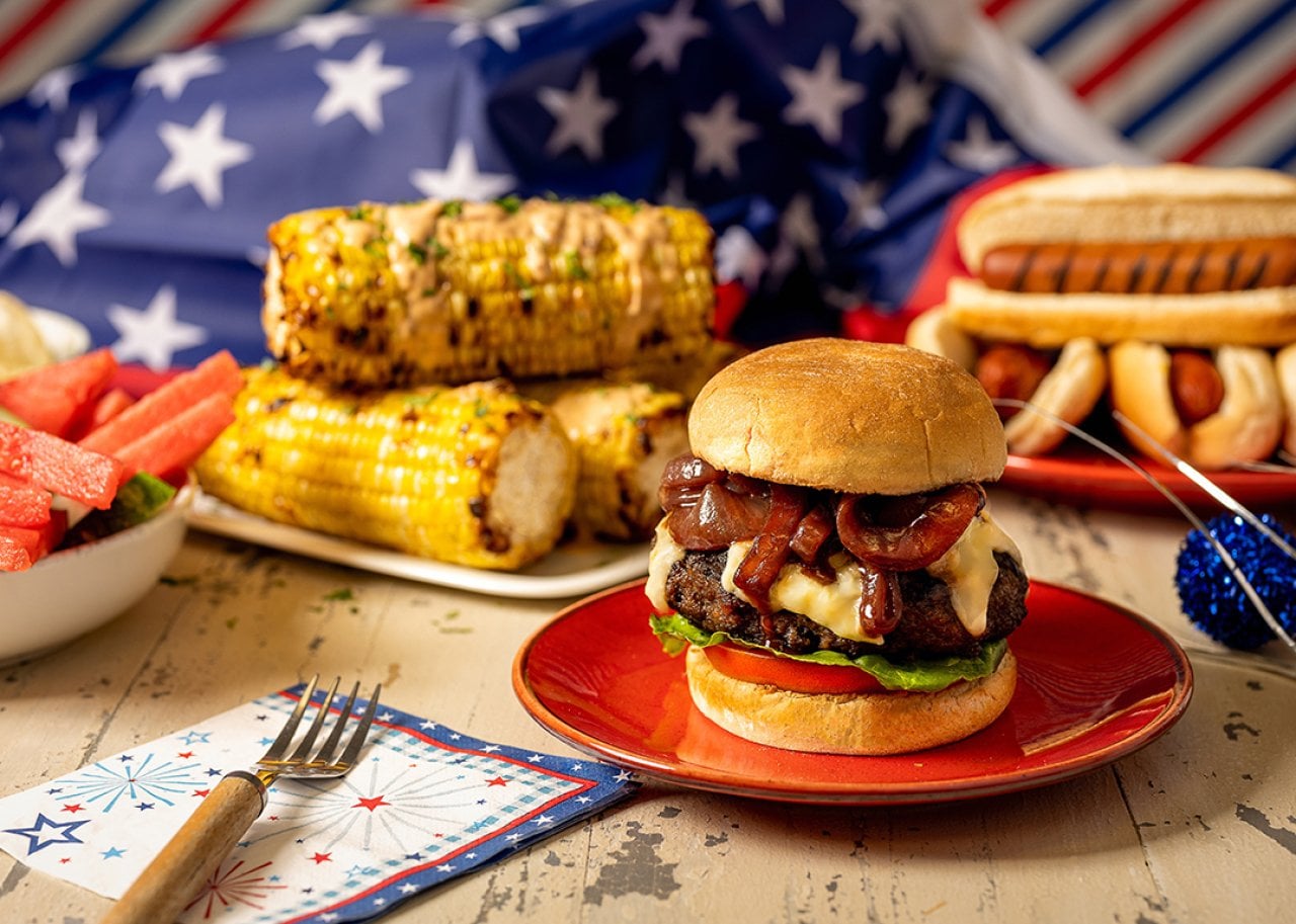 Cheeseburger with bacon and melted cheese on a bun, served on a red plate with corn on the cob and patriotic decorations in the background.