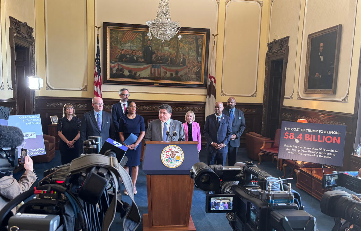 Politician at podium giving press conference with group of officials behind him in a formal government chamber, cameras recording nearby. Silence, flag and painting in background.