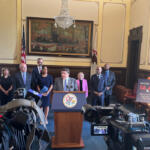 Politician at podium giving press conference with group of officials behind him in a formal government chamber, cameras recording nearby. Silence, flag and painting in background.