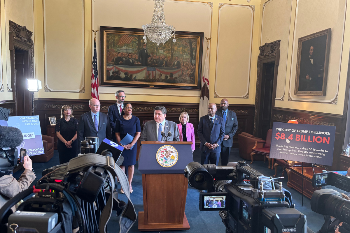 Politician at podium giving press conference with group of officials behind him in a formal government chamber, cameras recording nearby. Silence, flag and painting in background.