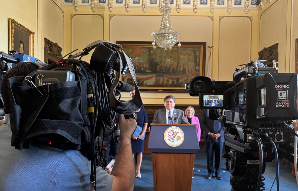 Politician at a podium delivering a press conference, surrounded by reporters and large broadcast cameras in an ornate room.