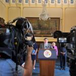Politician at a podium delivering a press conference, surrounded by reporters and large broadcast cameras in an ornate room.
