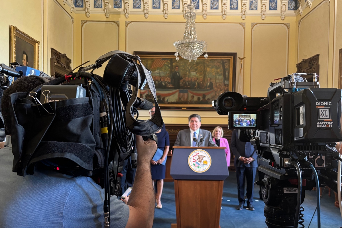 Politician at a podium delivering a press conference, surrounded by reporters and large broadcast cameras in an ornate room.