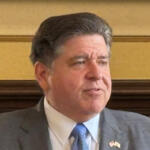 Middle-aged man in a gray suit and blue tie sits in a formal room, looking to the side.