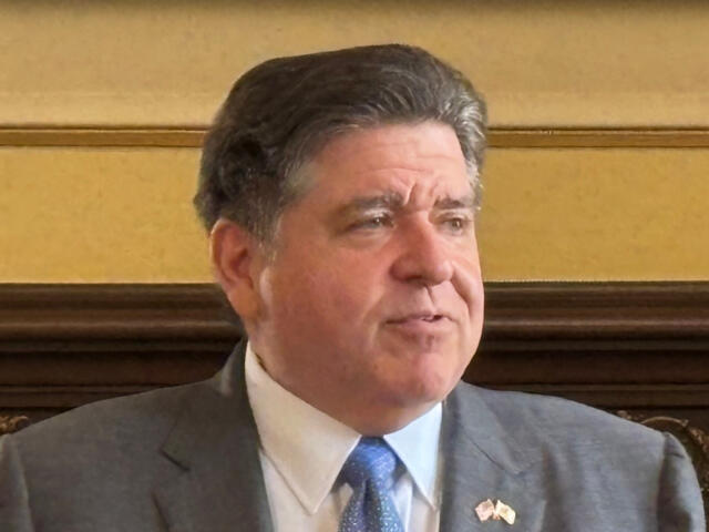 Middle-aged man in a gray suit and blue tie sits in a formal room, looking to the side.