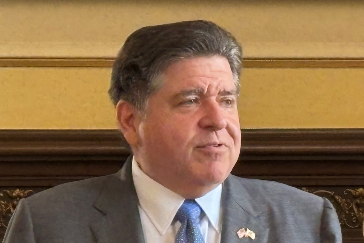 Middle-aged man in a gray suit and blue tie sits in a formal room, looking to the side.