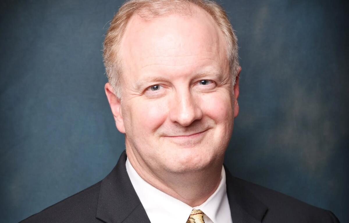Smiling man in a black suit and gold tie, head-and-shoulders portrait against a blue backdrop.