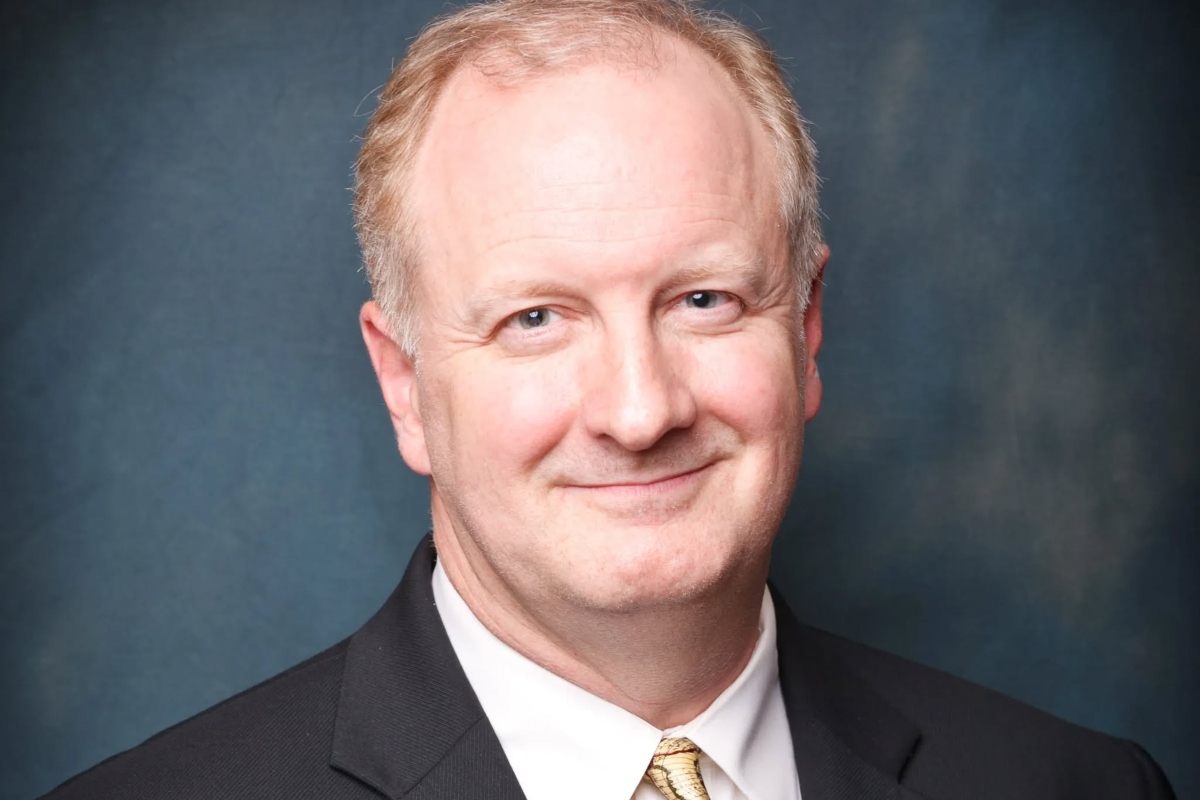 Smiling man in a black suit and gold tie, head-and-shoulders portrait against a blue backdrop.