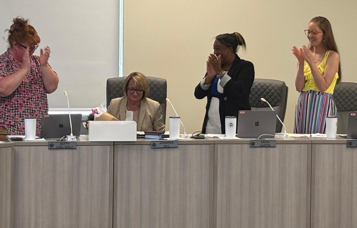 Four women in a meeting room applaud; one seated with a bouquet stands beside laptops, others stand and clap at the long conference table.